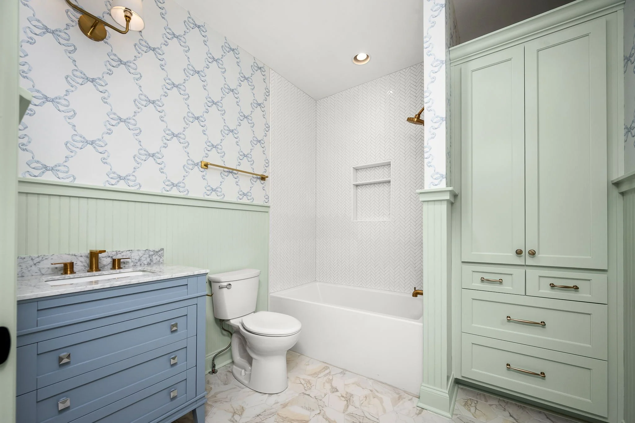 Bathroom with pastel green cabinetry, gold fixtures, with a patterned wallpapered wall and a marble countertop.