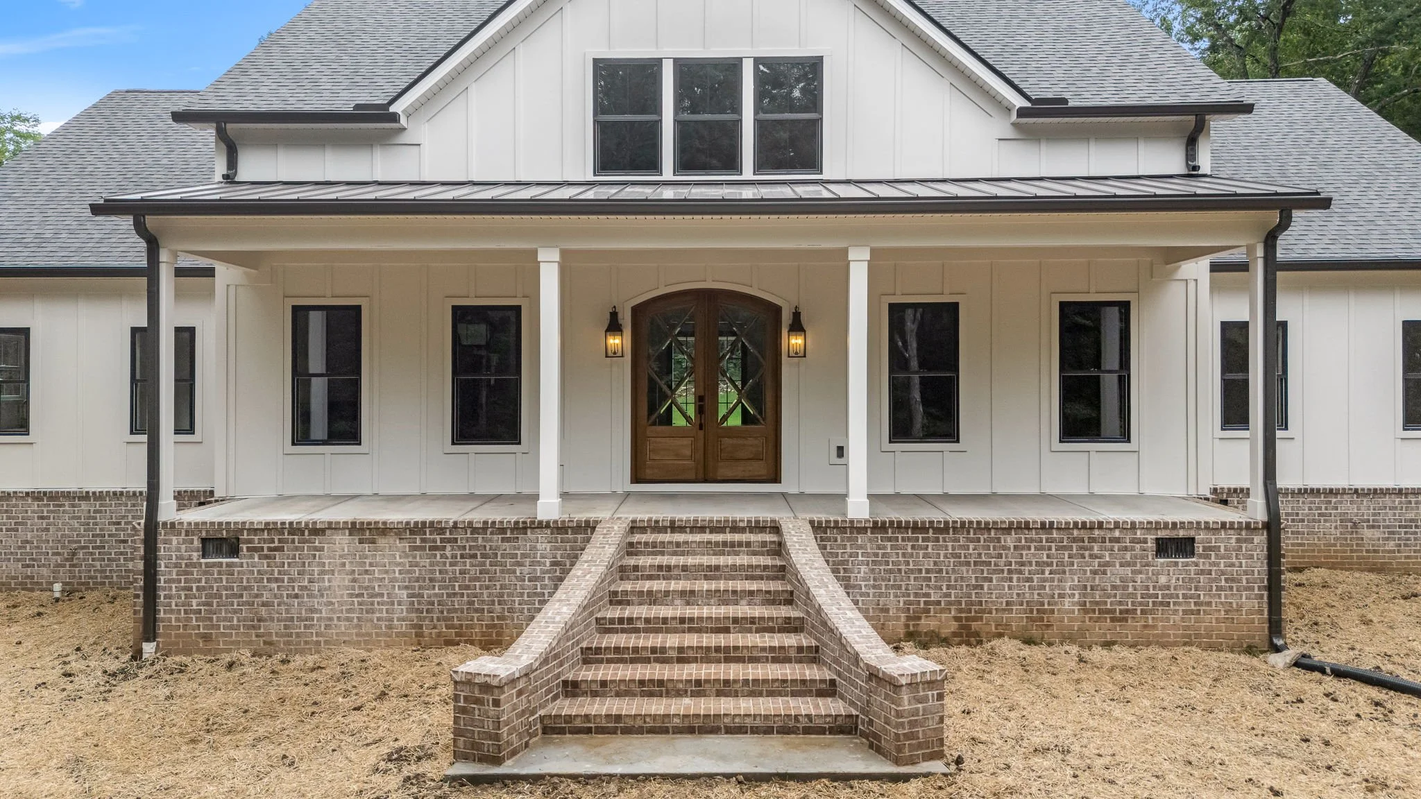 Front view of a modern house with a brick staircase leading to a porch with a wooden door, black-framed windows, white siding, and a gray metal roof, set against a partly cloudy sky.