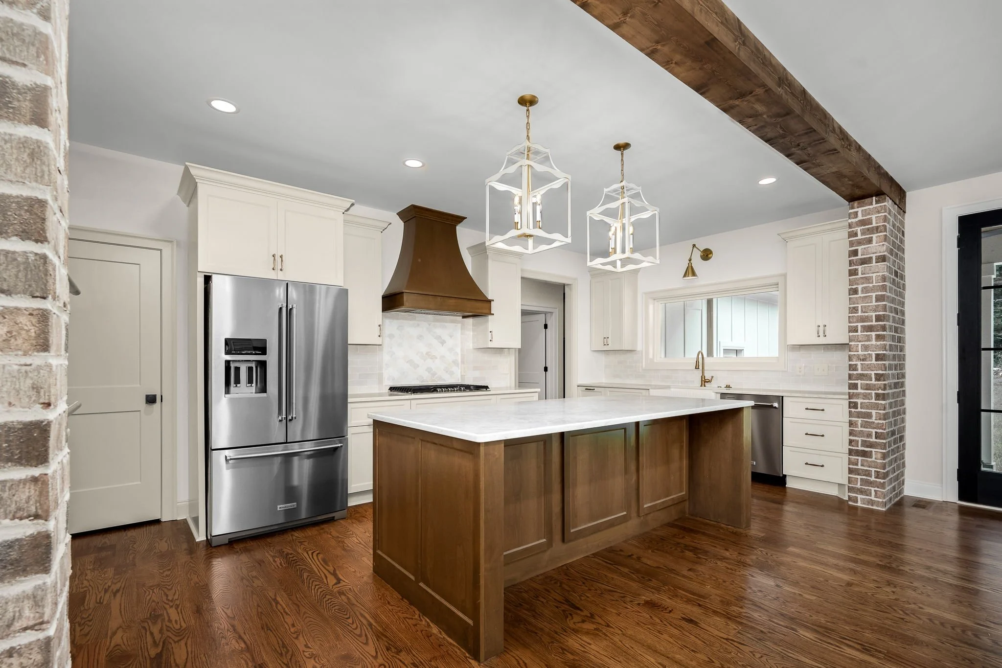 A kitchen with white cabinets, a stainless steel refrigerator, a wooden kitchen island, brick pillars, and modern pendant lights hanging from the ceiling.