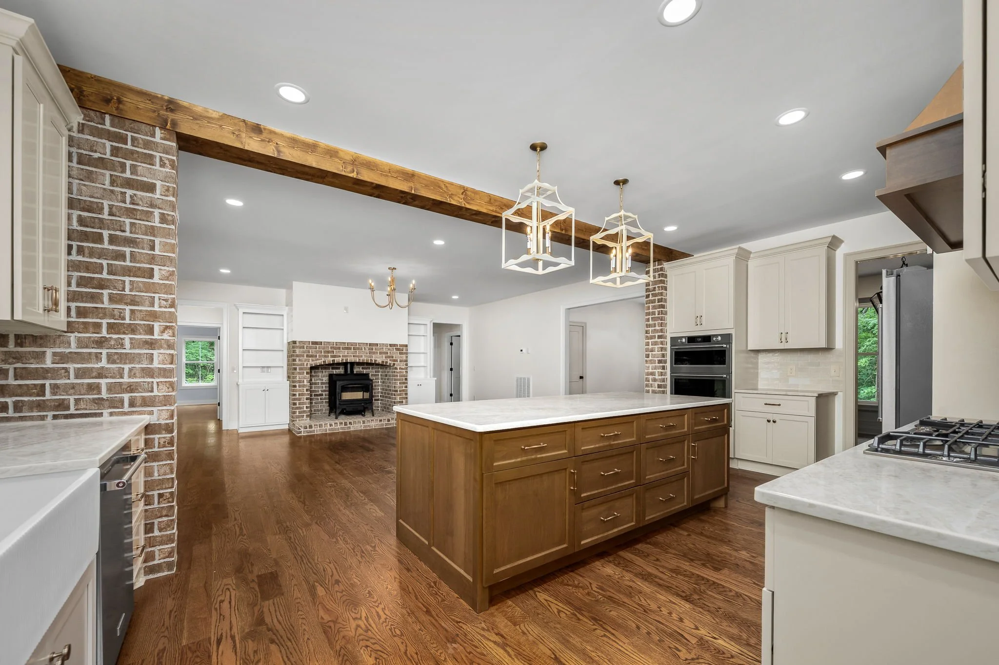 Interior view of a modern kitchen with a large island, wooden cabinetry, exposed brick walls, and a white ceiling with recessed lighting. Hanging light fixtures are also visible.