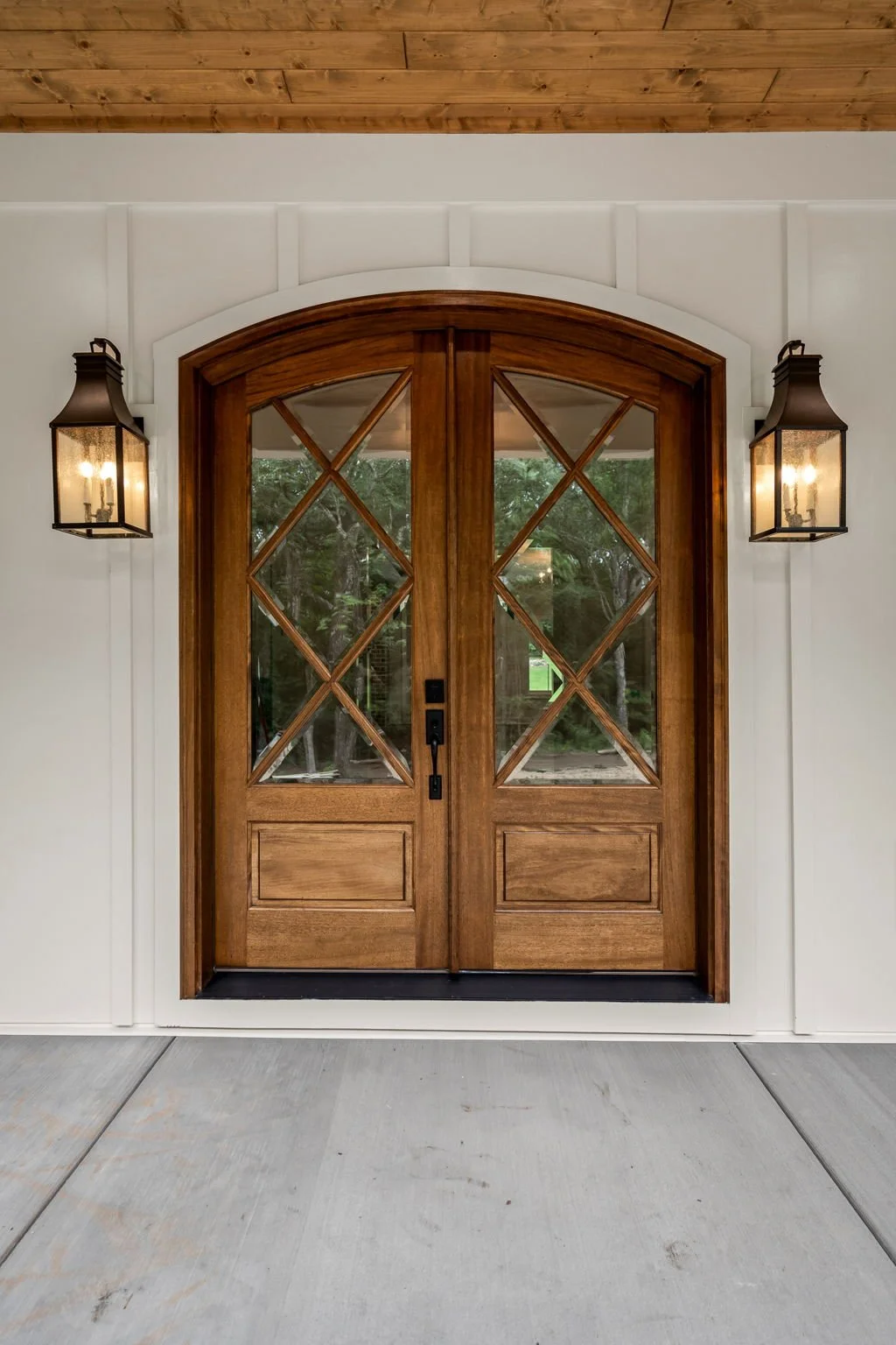 Double wooden front door with glass panels and decorative diamond pattern, flanked by black wall-mounted lanterns, set in a white wall with a wood-paneled ceiling above.