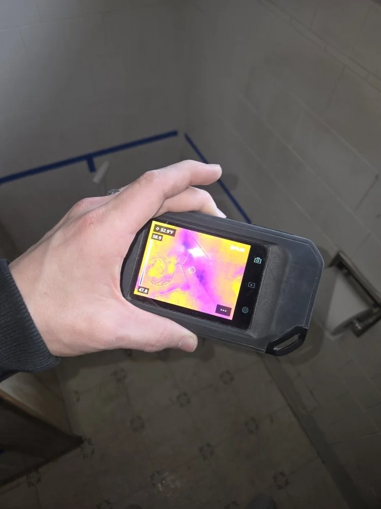 A hand uses a moisture meter finding water damage over a tiled bathroom floor, displaying a heat map on its screen.