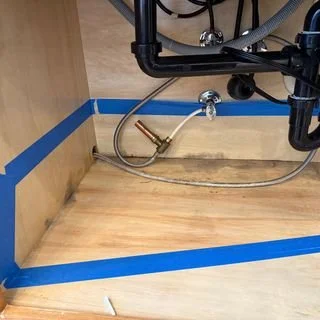 The inside of a cabinet under a sink in WA, with blue painters tape outlining the edges and exposed pipes visible.