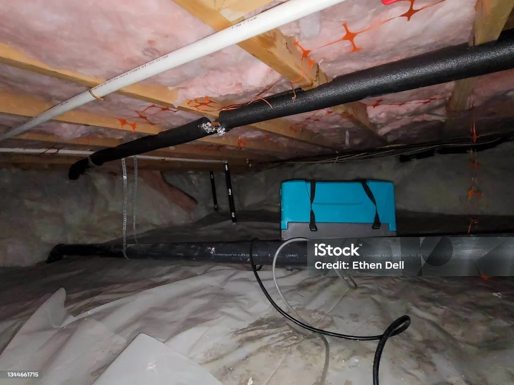 A crawl space with insulation, pipes, exposed beams, and crawlspace drying equipment removing moisture from the plastic-covered floor.