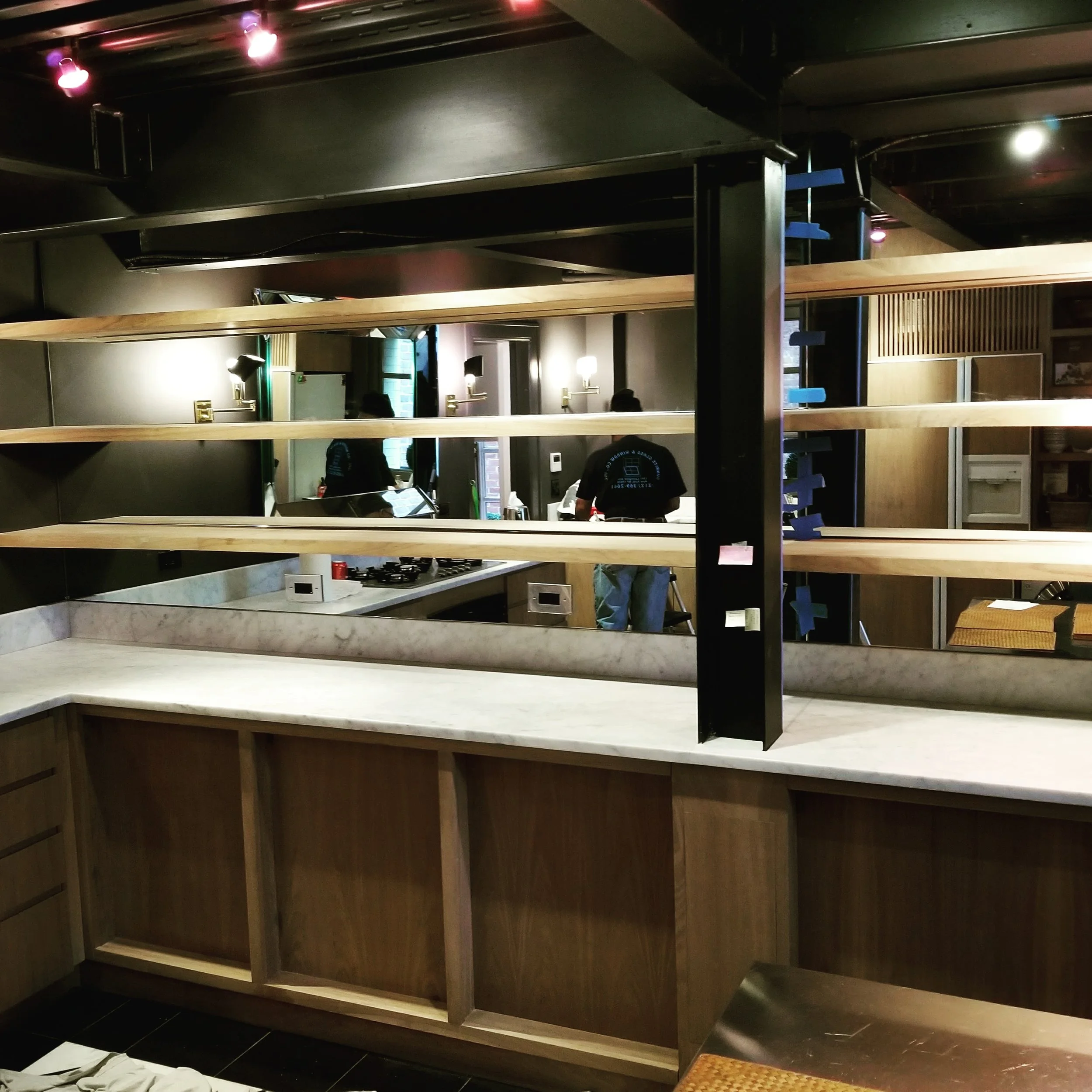 Interior view of a restaurant or cafe under construction with wooden shelves, marble countertops, and workers seen in the reflection of a mirror.