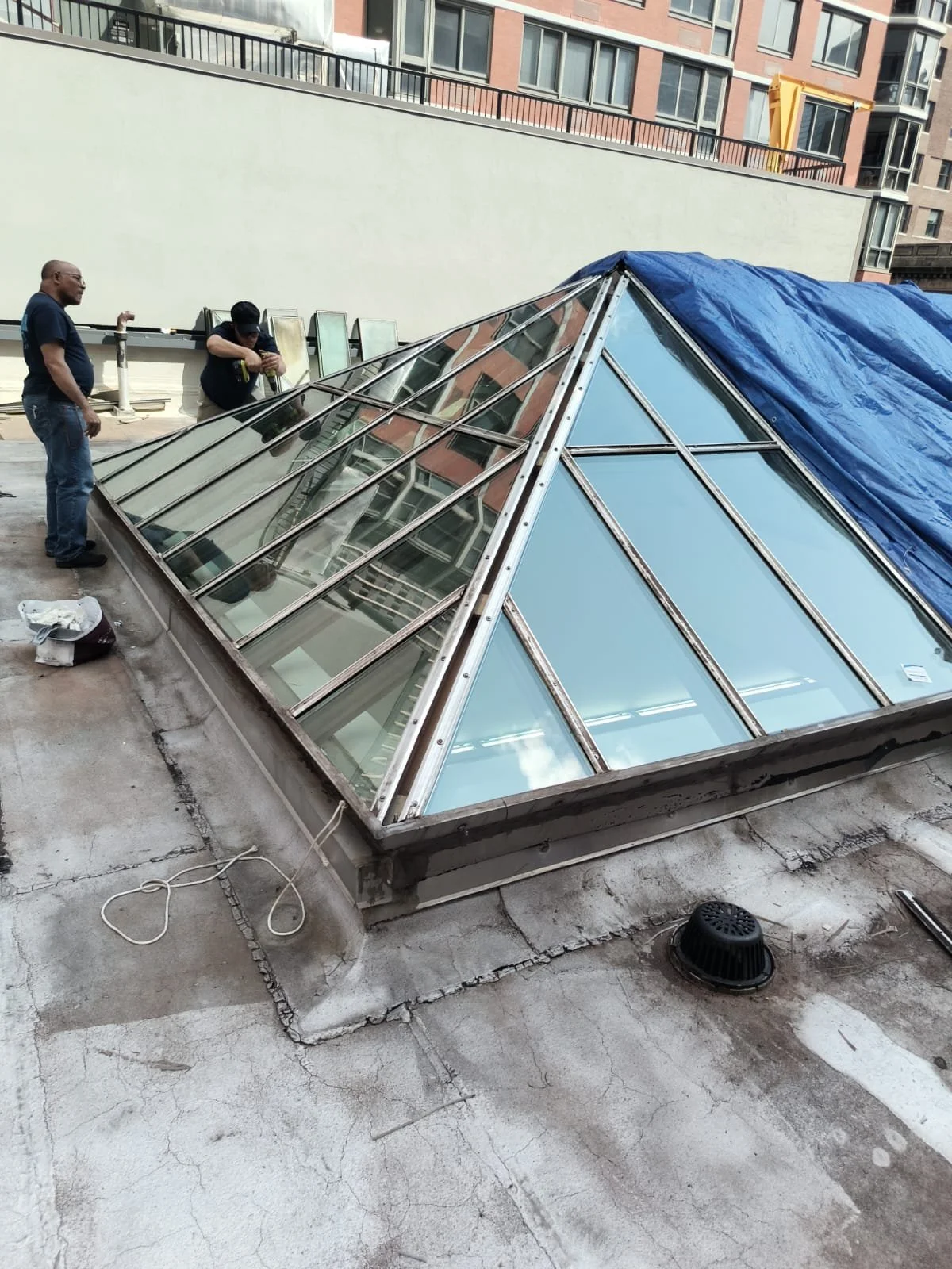 A rooftop skylight with a metal frame and glass panels, partially covered by a blue tarp. Two workers and one person are on the roof near the skylight, working or inspecting it. Buildings are visible in the background.