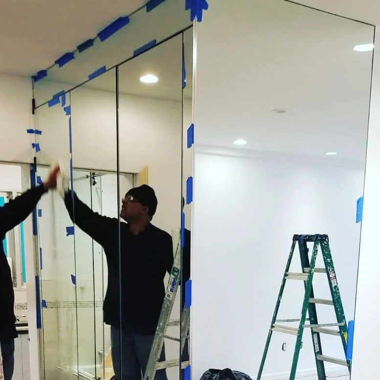 Workers installing a mirrored wall in a room, with ladders and blue painter's tape around the edges of the mirrors.