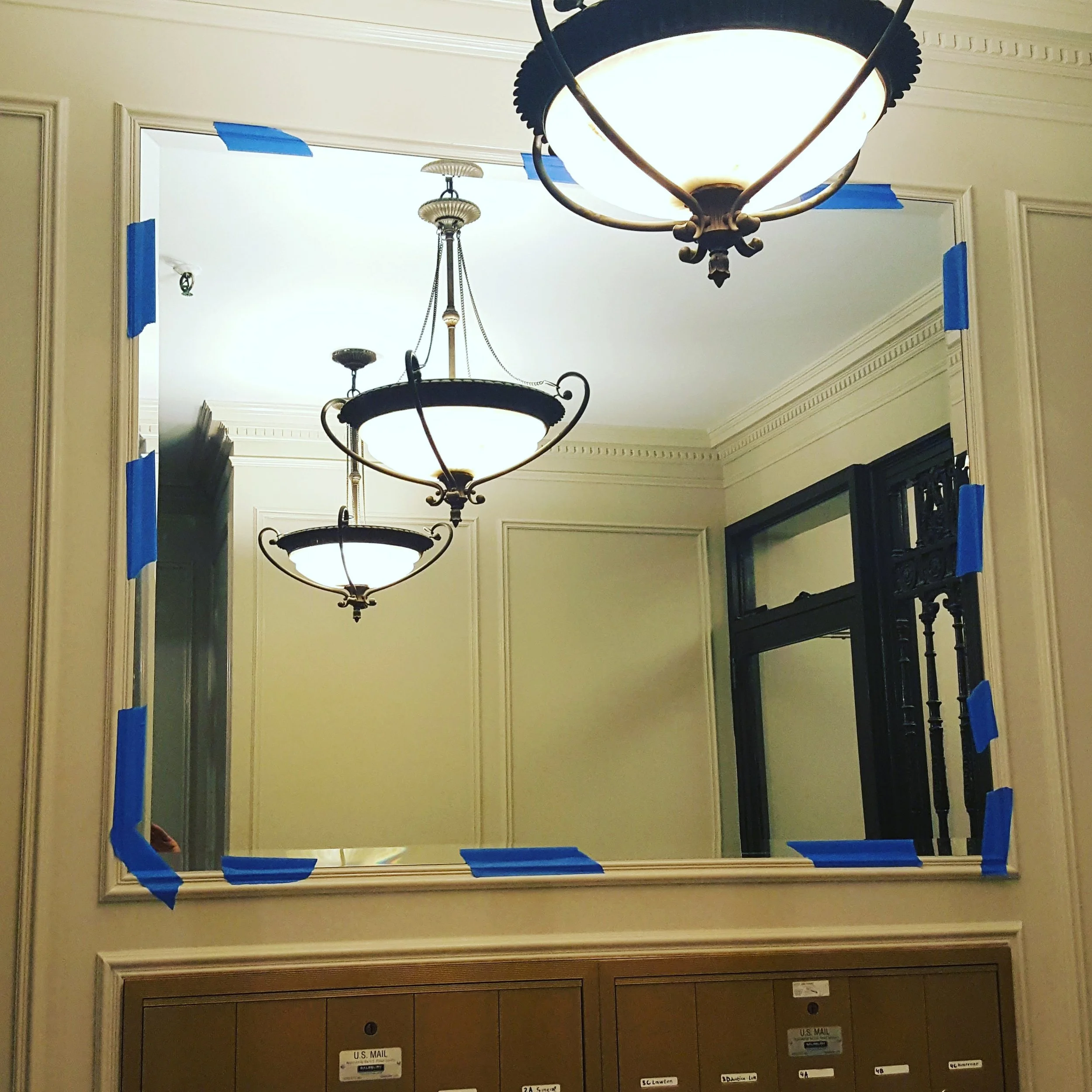 A large mirror with blue painter's tape around the edges hangs on a beige wall, reflecting three ornate ceiling chandeliers in a well-lit hallway with crown molding.
