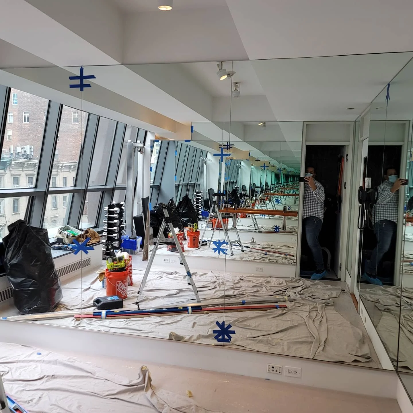A room with large angled windows, a wall of mirrors, and construction tools and supplies on the floor. The ceiling has small adjustable lights, and a person wearing a mask, plaid shirt, and jeans is taking a photo of the room's reflection.