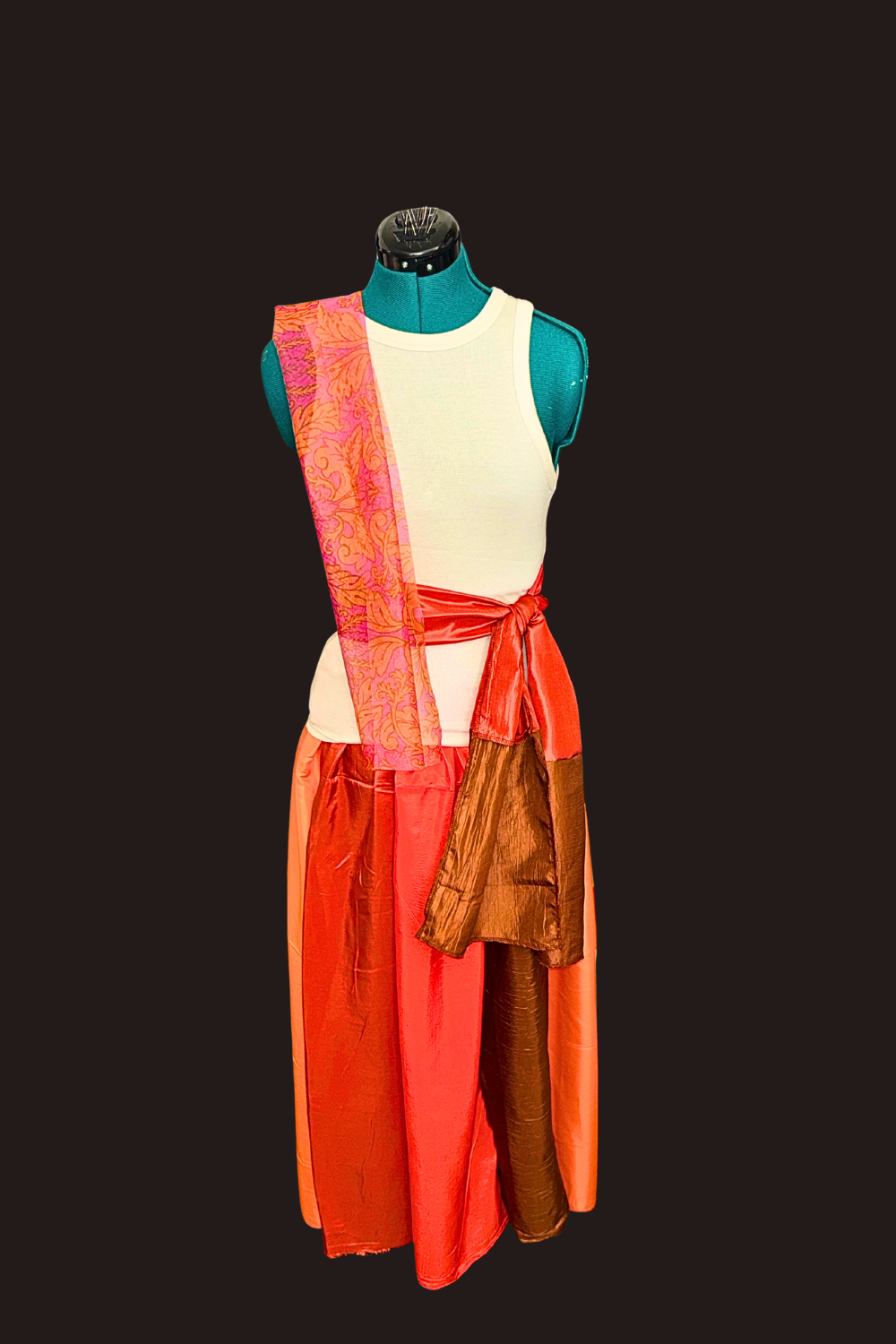 A mannequin dressed in a sleeveless white top with an orange wraparound sash, red, orange, and brown flowing skirt, cream tank top, orange & red dancing scarves, set against a dark background.