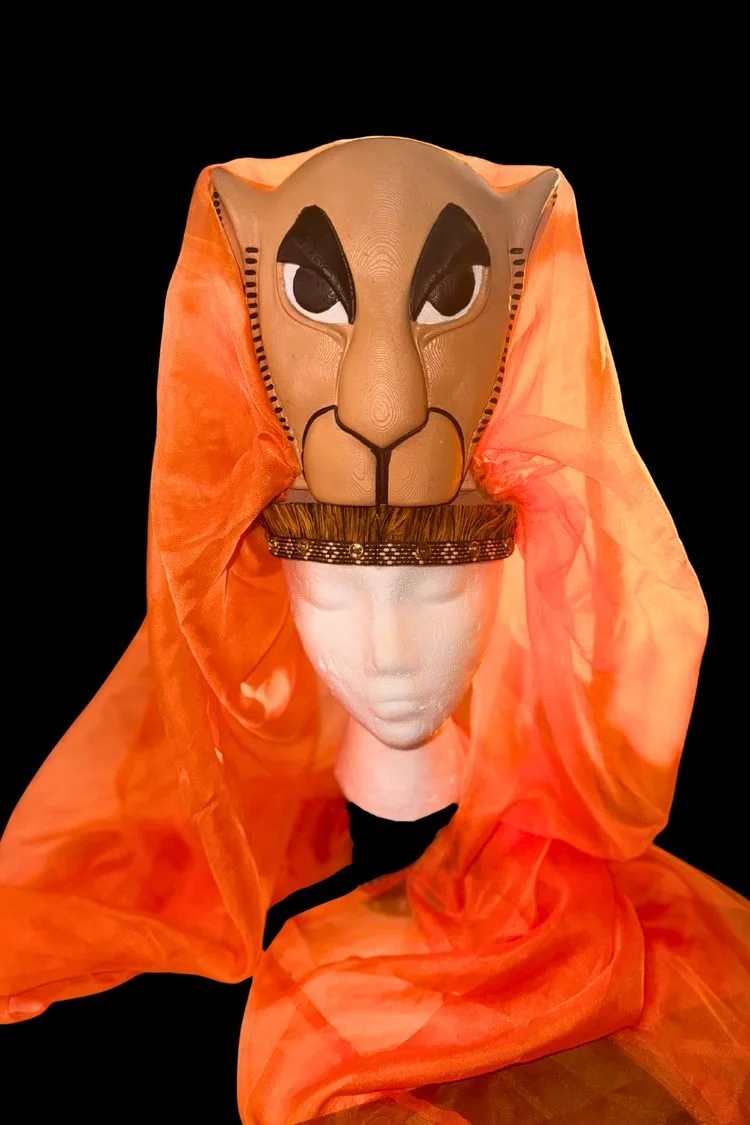 Handmade lion face headpiece featuring tan and orange facial details, dark eyes, and flowing orange fabric, displayed on a mannequin for The Lion King JR. school musical.