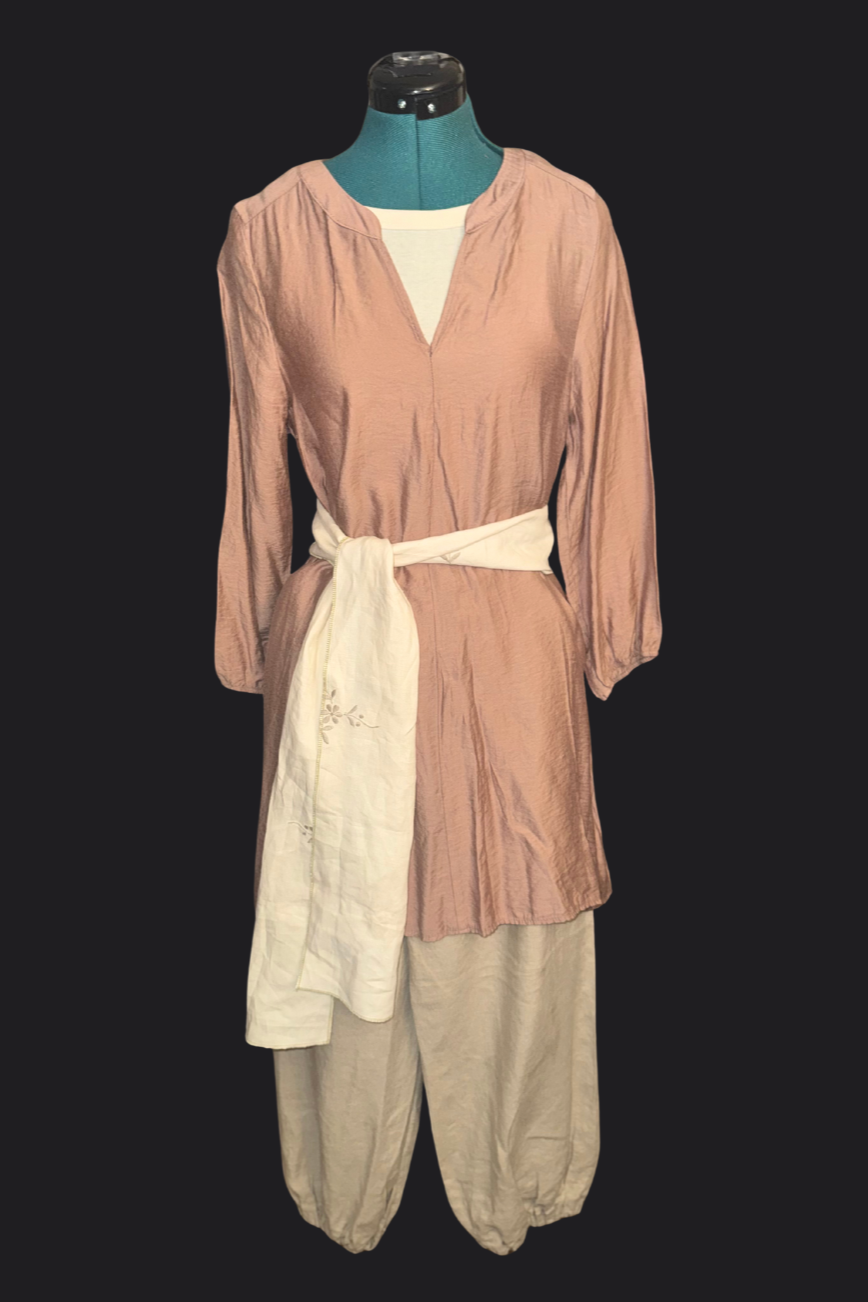 A mannequin dressed in a rust long-sleeve dress with a cream sash at the waist, paired with loose beige pants. The background is black.