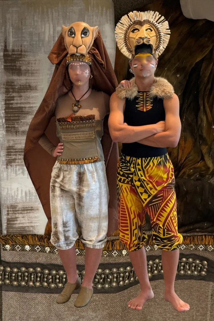 Two performers wearing lion masks, patterned costumes, and colorful African-inspired fabrics during a stage production of Disney's The Lion King JR.