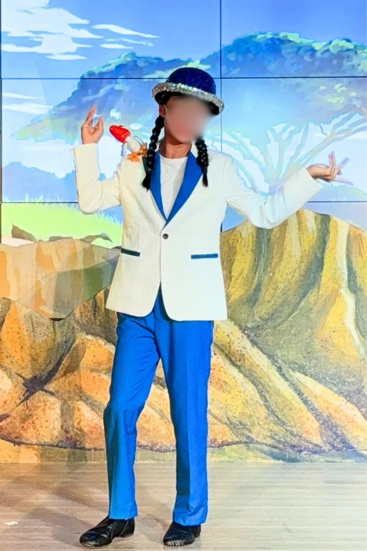 Lion King JR. Zazu Costume: Young performer wearing a blue-and-white bird-inspired costume with a white jacket, blue pants, and coordinating blue hat, posing in front of a painted landscape backdrop for The Lion King JR.