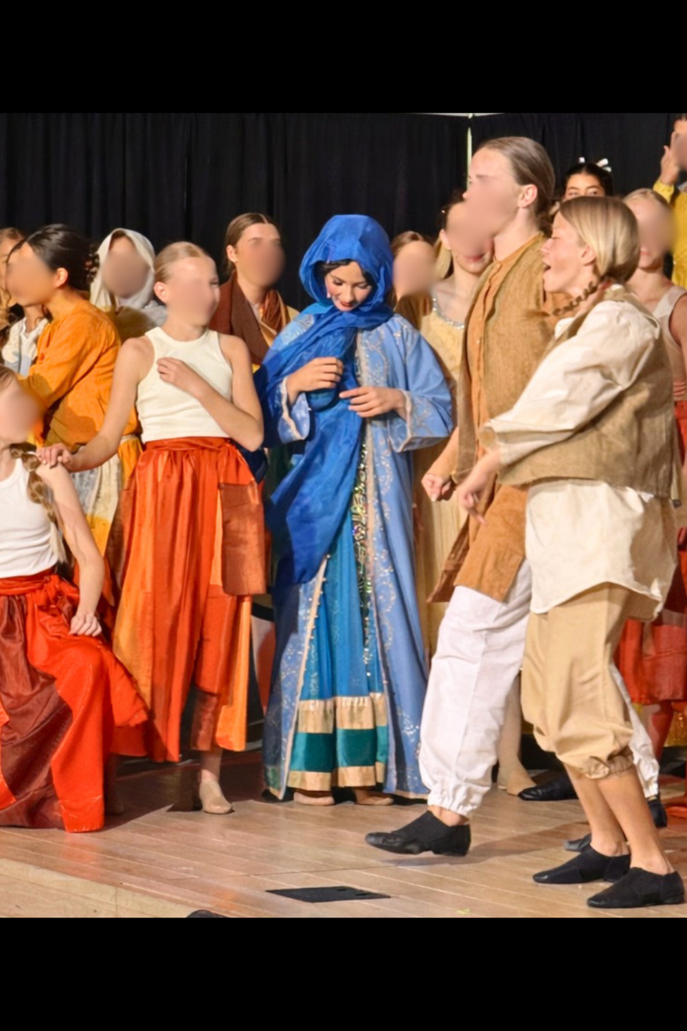 Princess Jasmine in disguise at the Agrabah market in Aladdin Jr, wearing a blue cloak among dancing townspeople during a school musical scene.
