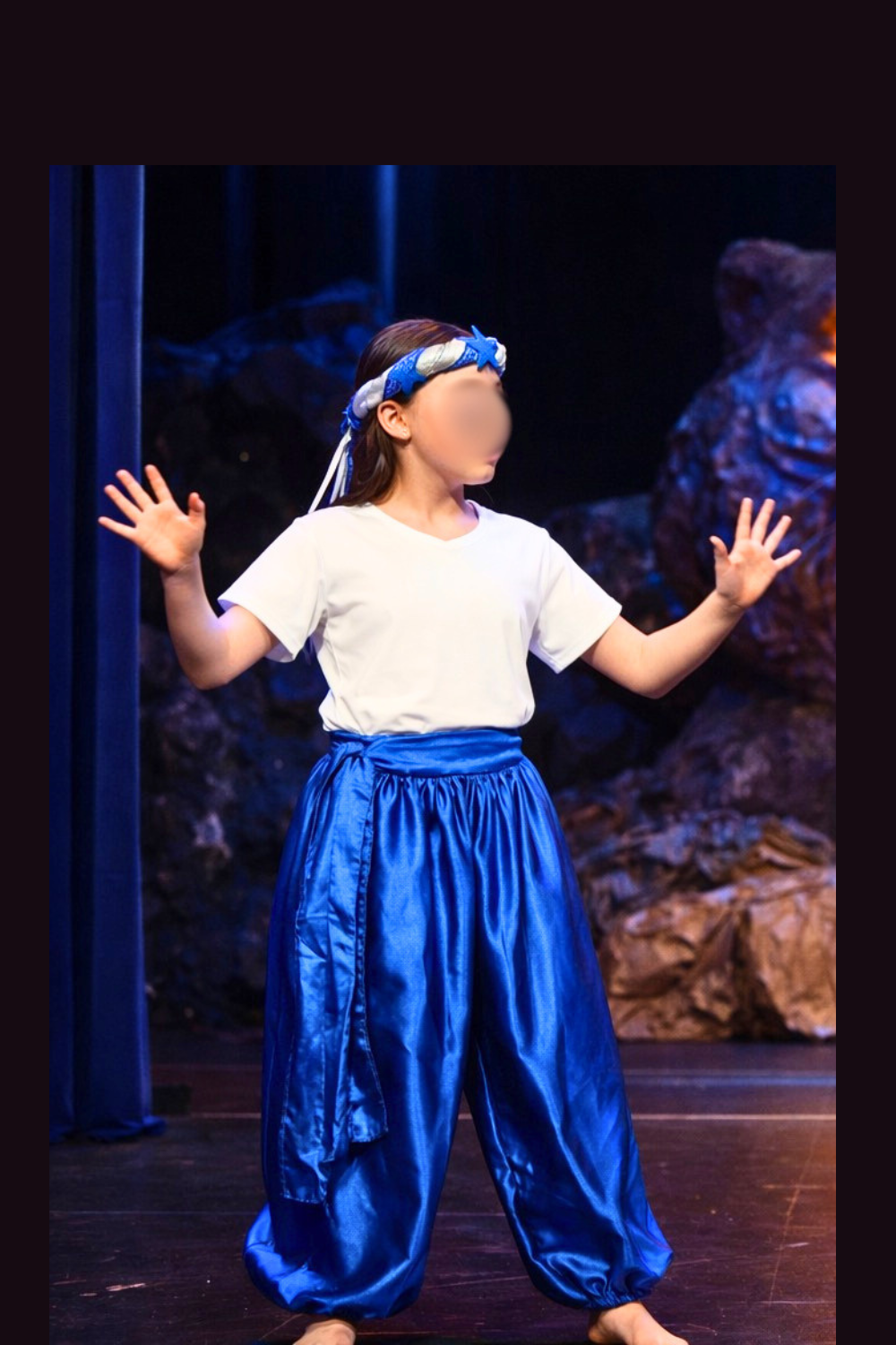 A young girl performing on stage, genie assistants in the Cave of Wonders scene in Aladdin Jr wearing blue satin pants, white tops, and matching braided headbands during a school musical performance.