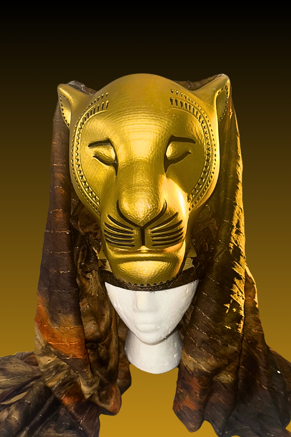 A golden lion face mask mounted on a white mannequin head, draped with a brown, textured cloth with a burnt look.