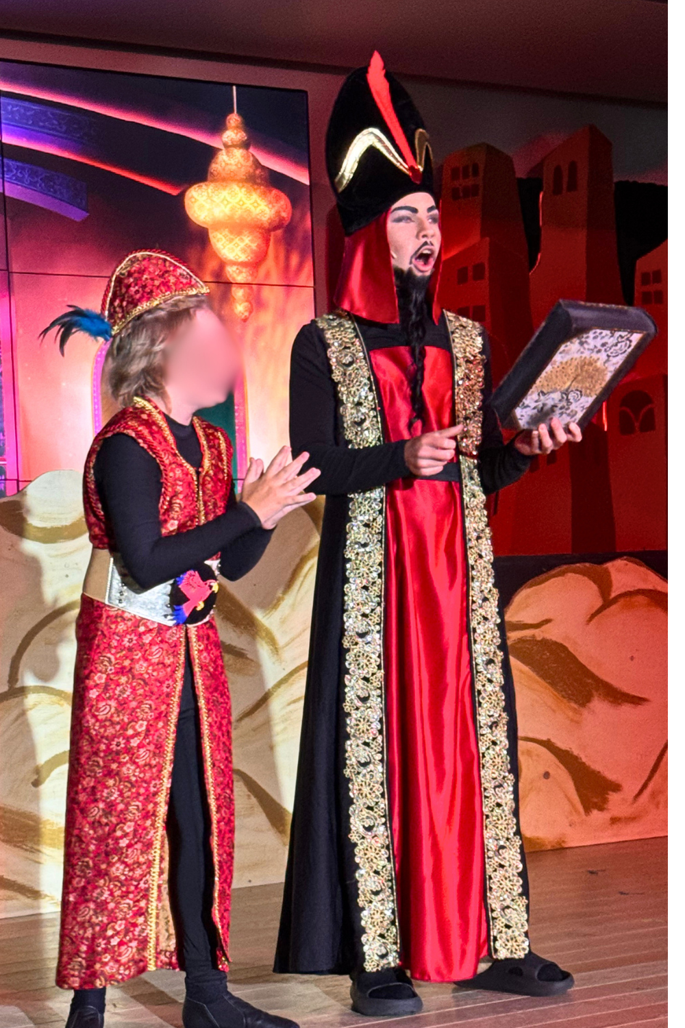Jafar and Iago on stage in Aladdin Jr, wearing black and red robe with gold trim and red brocade vest during a school musical performance scene.