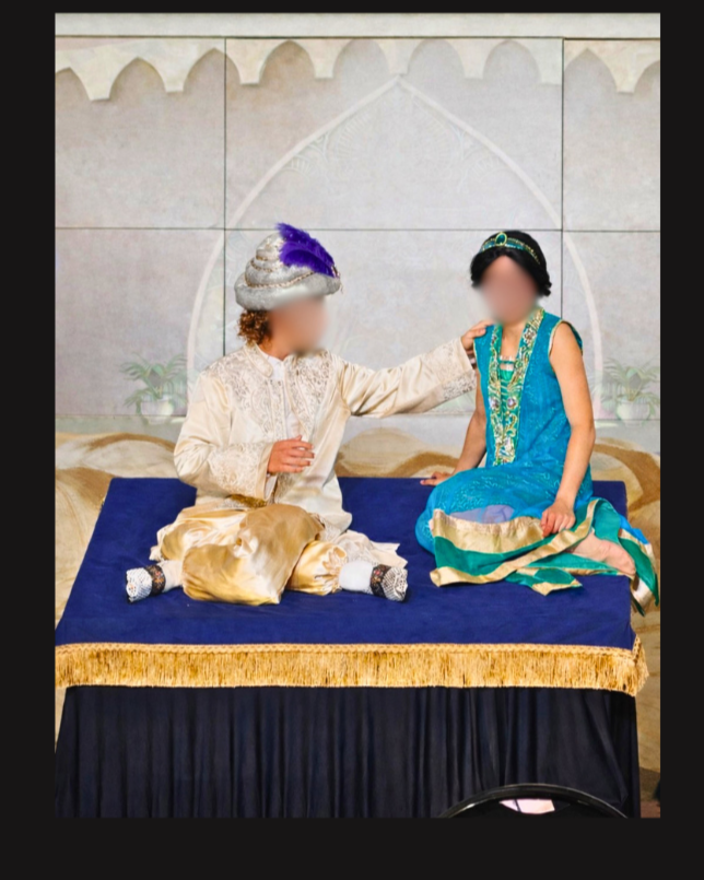 Aladdin and Princess Jasmine seated on a rolling magic carpet platform in Aladdin Jr, wearing cream and turquoise costumes during a school musical scene.