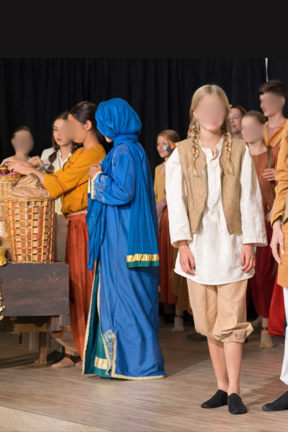 Children dressed in costumes, participating in a play or reenactment, standing on a stage with a black curtain backdrop. Set in Agrabah Market in Aladdin Musical