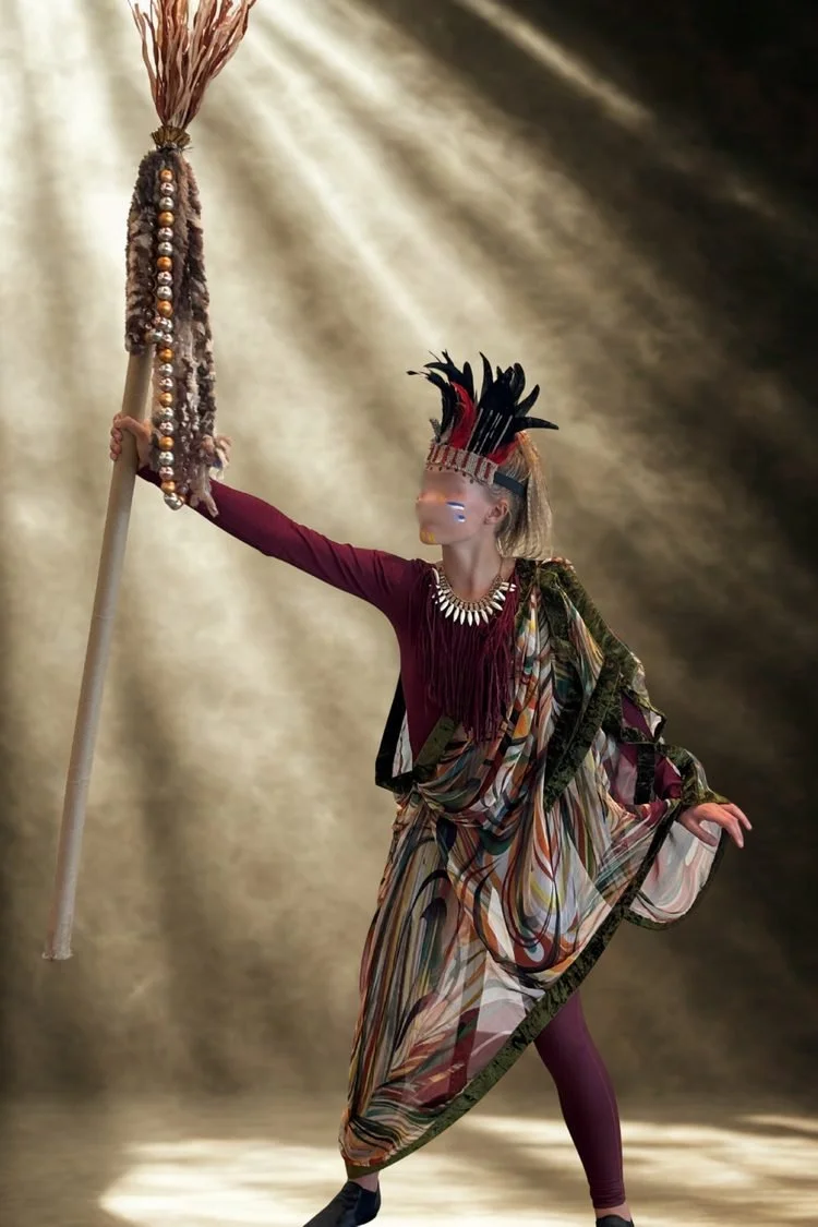 Performer portraying Rafiki in Disney's The Lion King JR. wearing a dramatic feathered headdress, red and black costume, and decorative necklace while holding a tall staff during a theatrical performance.