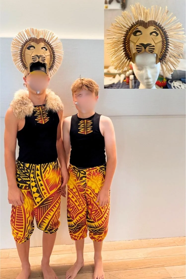 Two young performers wearing coordinated black tops and colorful African-inspired patterned pants. One wears a handmade lion mask with a straw-like mane and feathered collar for Disney's The Lion King JR.