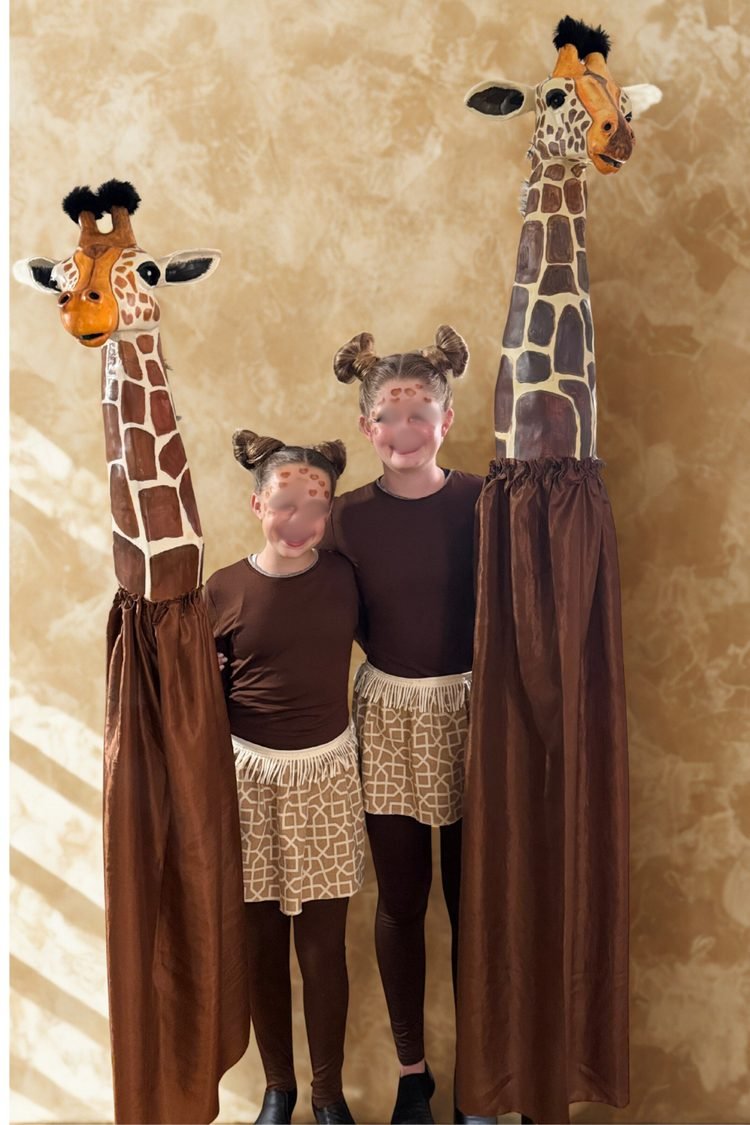 Lion King JR – Giraffe Costume: Two young performers wearing brown and African-print costumes standing beside tall handmade giraffe puppets with long necks and spotted faces for The Lion King JR.