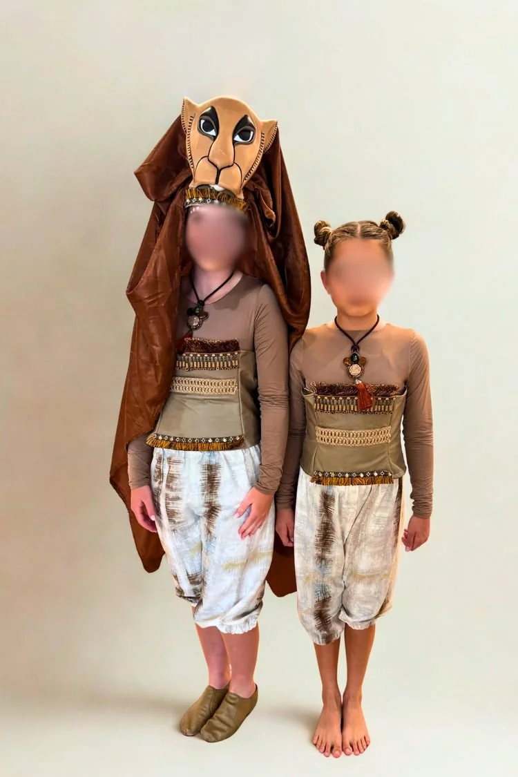 Two young performers modeling Lion King JR. costumes featuring lion-inspired headpieces, patterned tunics, loose white pants, and coordinated accessories for a school theater production.