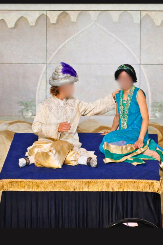 Two children dressed in traditional attire sitting on a stage with a backdrop resembling Indian architecture. The child on the left, in cream-colored clothing with a turban and purple feather, touches the shoulder of the child on the right, who is dressed in blue and green traditional clothing with a headpiece.