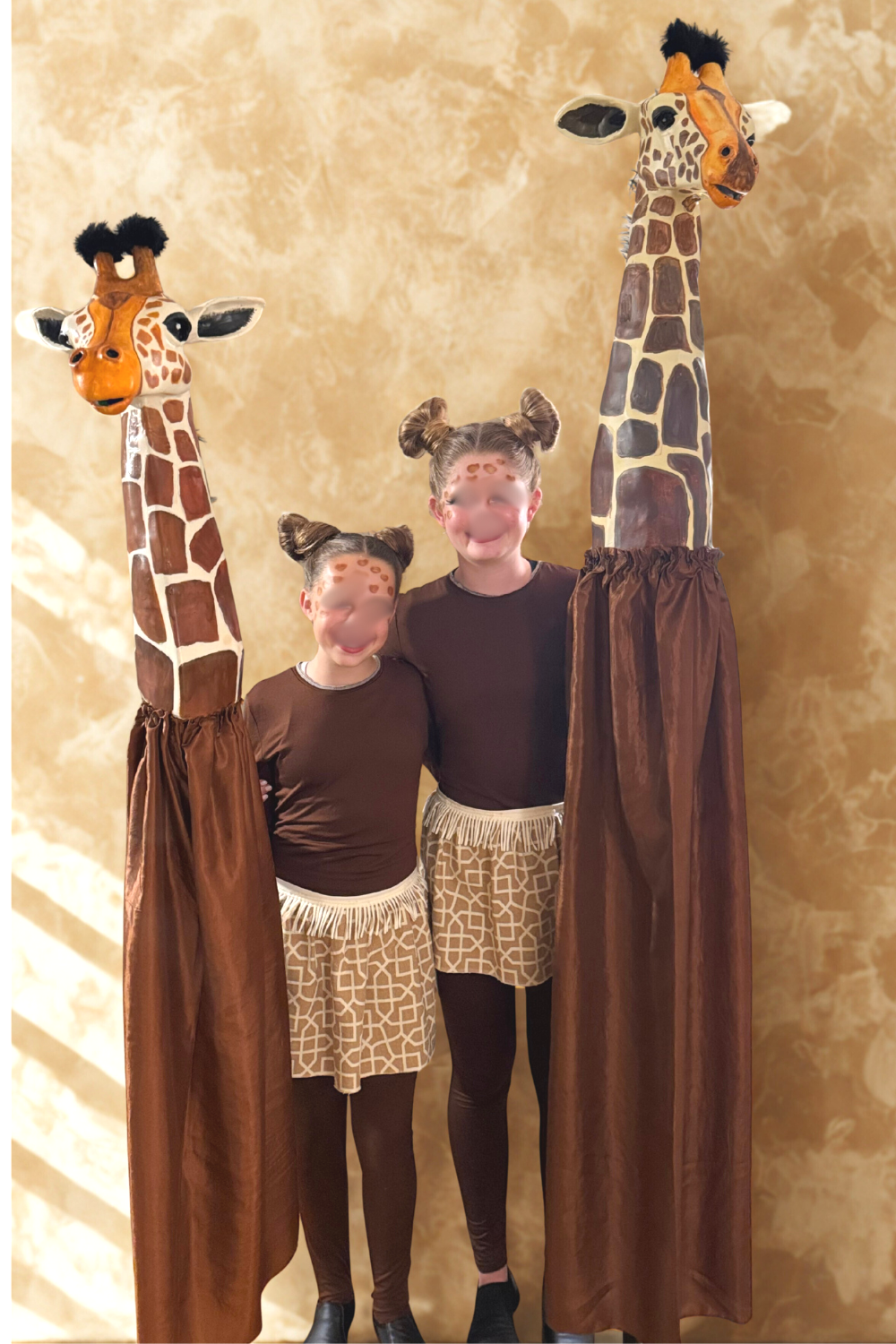 Two children dressed as giraffes standing between two giraffe puppets with long necks, ears, and spots, against a beige background.