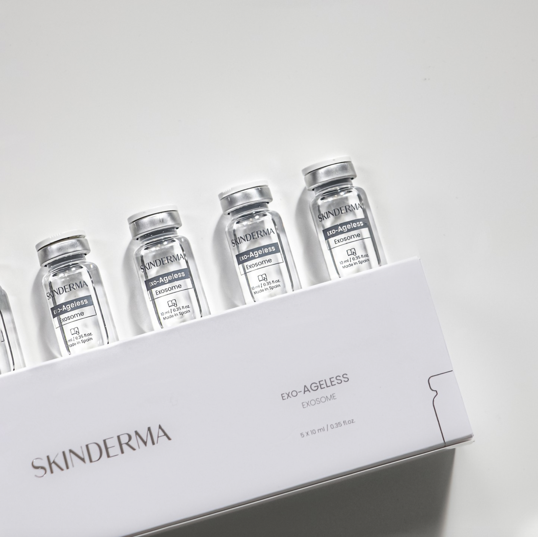 Een kleine fles met Skinderma Exo-Ageless Exosome serum op een witte ondergrond, met een medische machine en een witte box met het woord 'SKINDERMA' op de achtergrond.