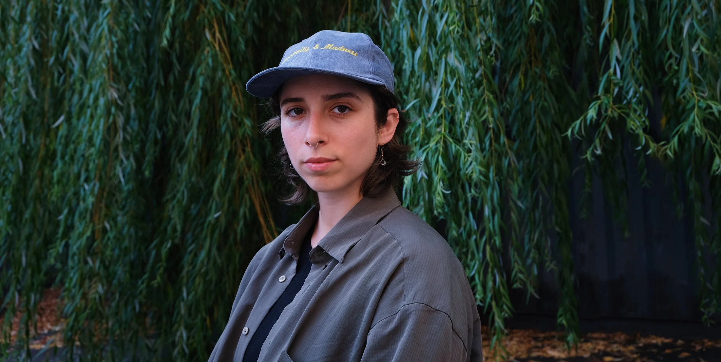 A young person with short dark hair, wearing a blue baseball cap with yellow embroidery, a gray button-up shirt over a black top, and earrings, standing in front of green leafy trees.