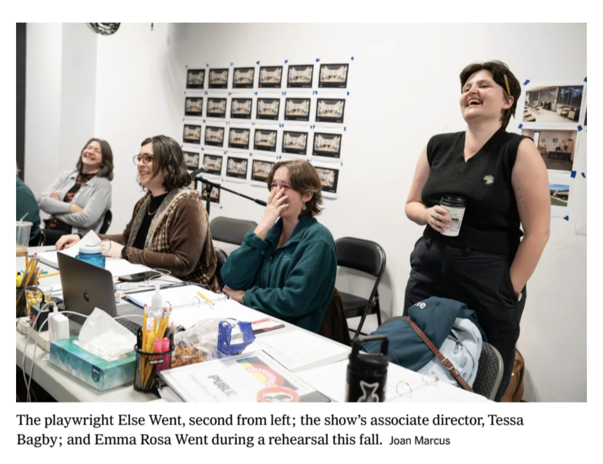 A group of four women sitting at a table, laughing and smiling during a rehearsal. One woman is standing, holding a coffee cup, and laughing. The table has various items including a laptop, tissues, snacks, and papers.