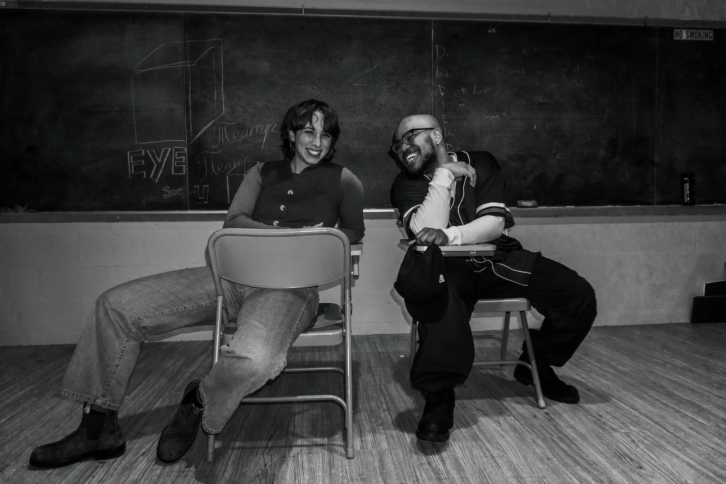 Two theater artists sitting on chairs in a classroom, laughing and smiling at each other, with a blackboard behind them.