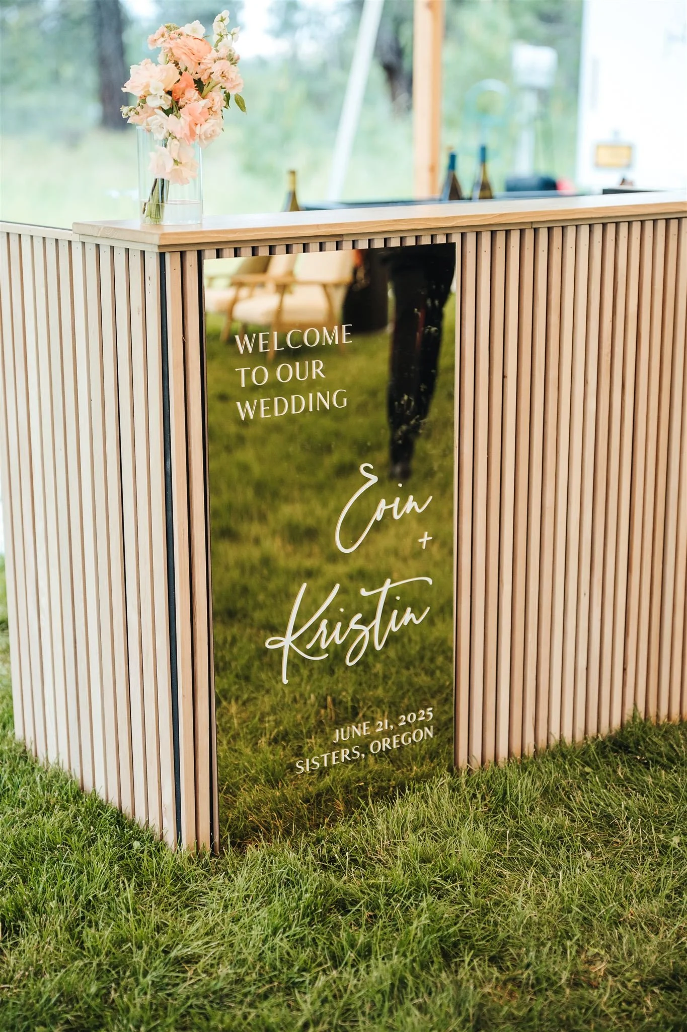 A wooden welcome sign at a wedding, with the text "Welcome to our wedding Erin + Kristin June 21, 2025 Sisters, Oregon". The sign features a background image of a wedding couple and is decorated with a bouquet of light pink flowers in a clear vase on top.