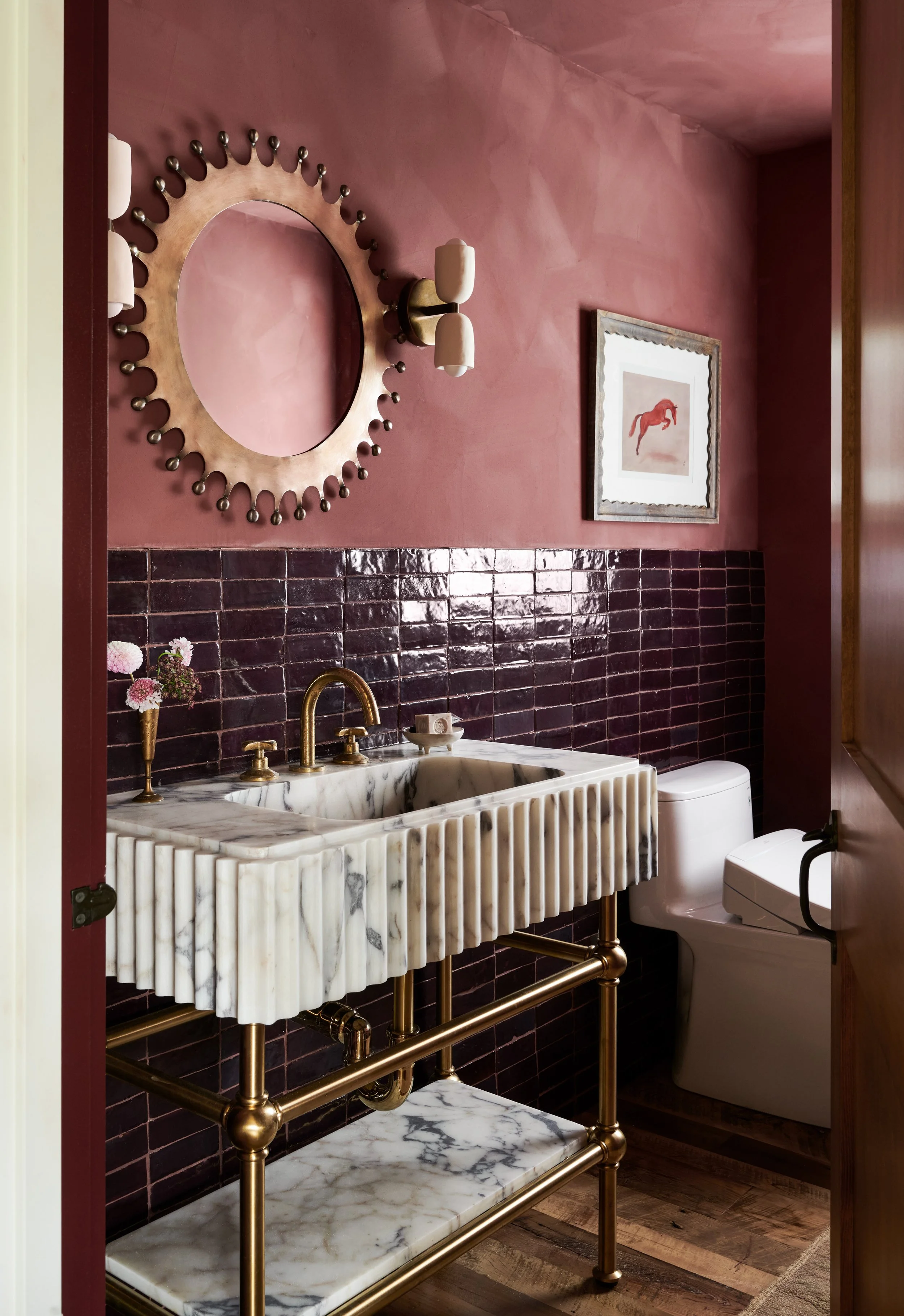 Powder room with rose plaster walls, aubergine tile, fluted marble console sink on brass legs and sunburst mirror by Ivory & Bone Interiors, Groveland, MA