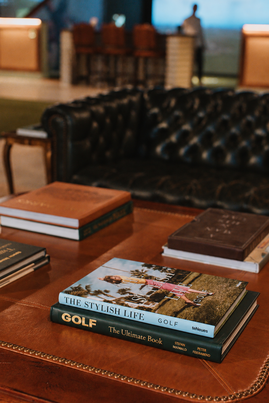 Golf books on a leather ottoman with a tufted Chesterfield sofa at Coach Club golf lounge in Newburyport, MA, designed by Ivory & Bone Interiors