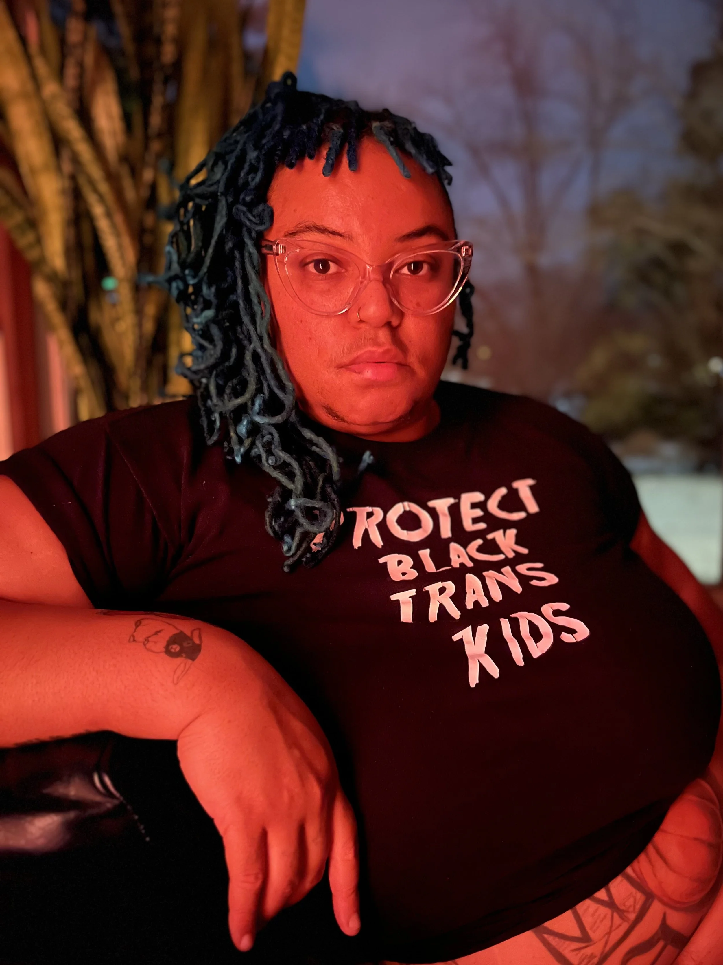 A fat lightskinned Black person with a round face, turquoise locks and bangs, cat eye glasses, tattoos, and nose ring sits sideways under orange light wearing a ‘protect Black trans children’ shirt; snowy scene behind.