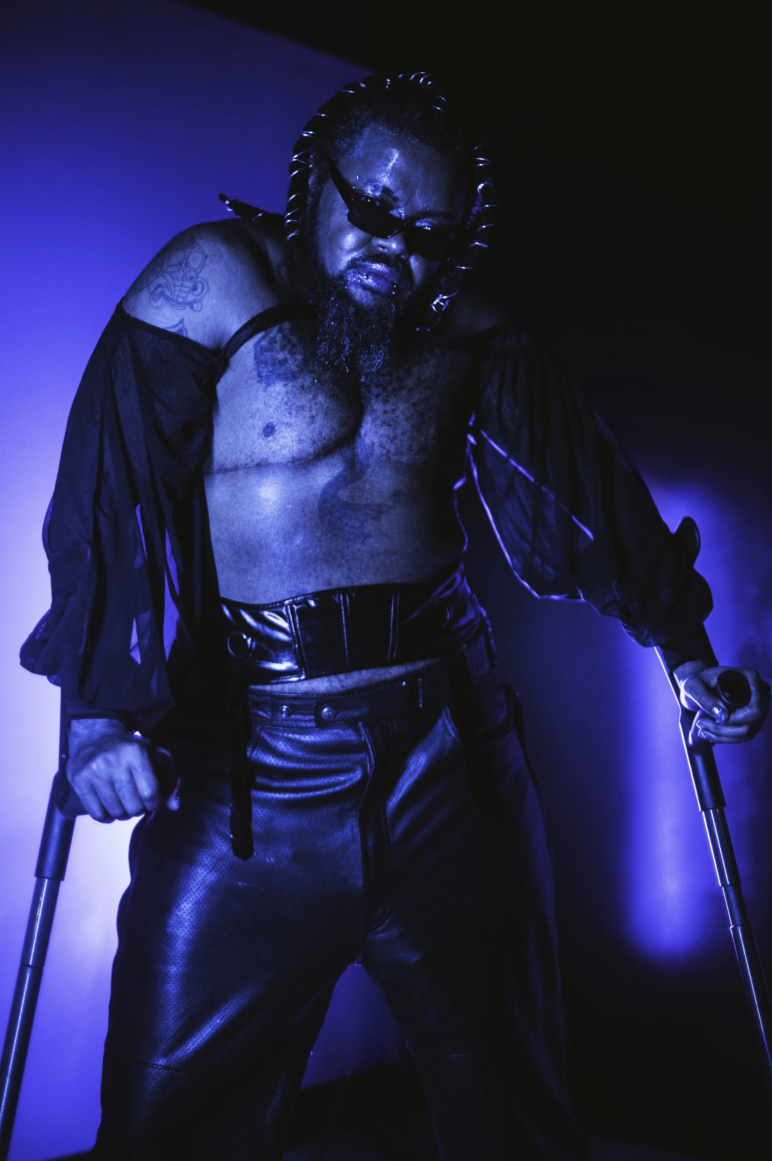 Uhuru is standing in a blue-lit room, wearing black mesh sleeves, his chest out, leather pants, and a corset, with his arm crutches. He has thick locs with silver wrap on them, and sunglasses. Photo by Rey Broughton.