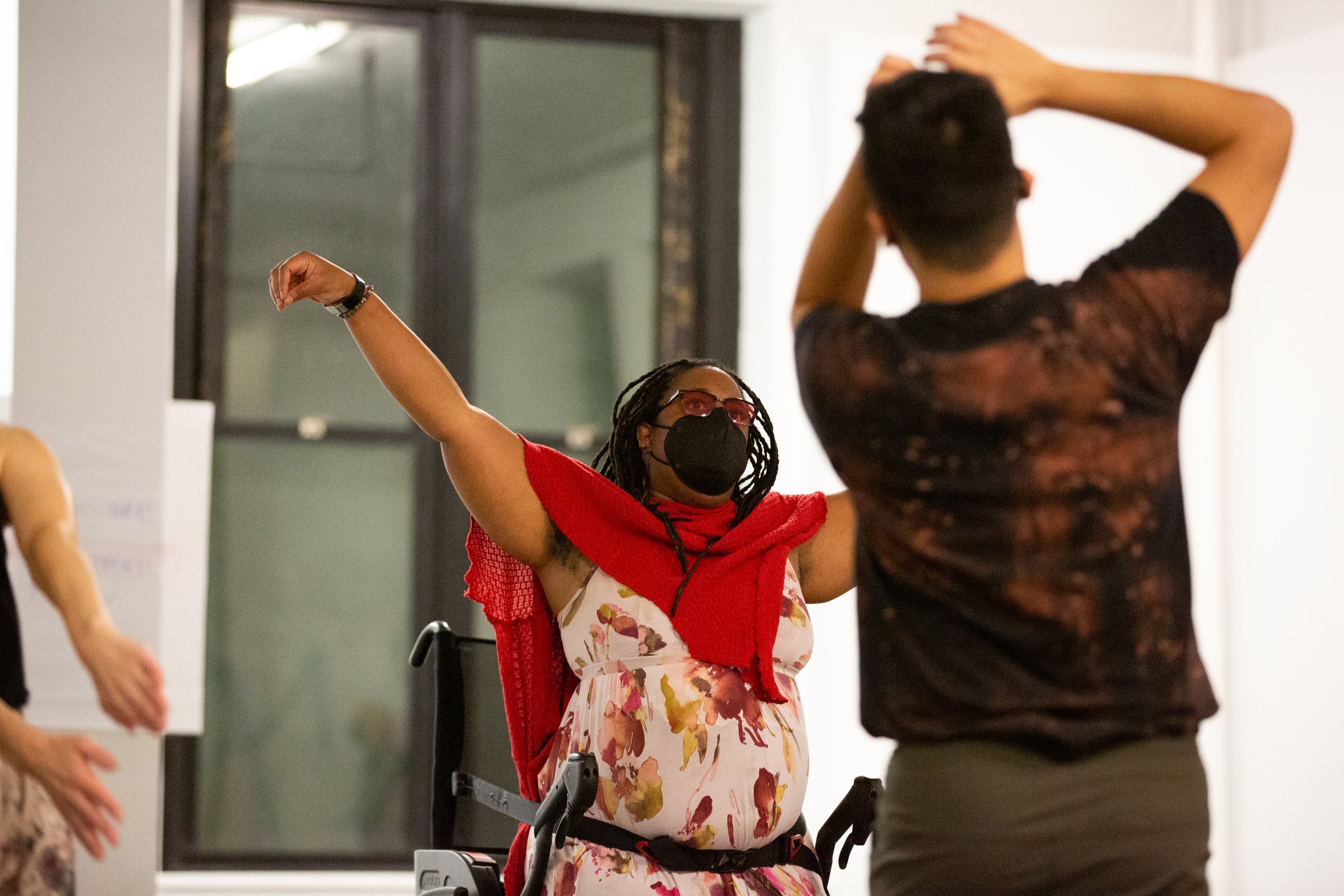 Two people dancing, one Black woman and one white man. Both have their arms outstretched. The woman is using an assistive wheelchair, the man in standing and seen from behind.