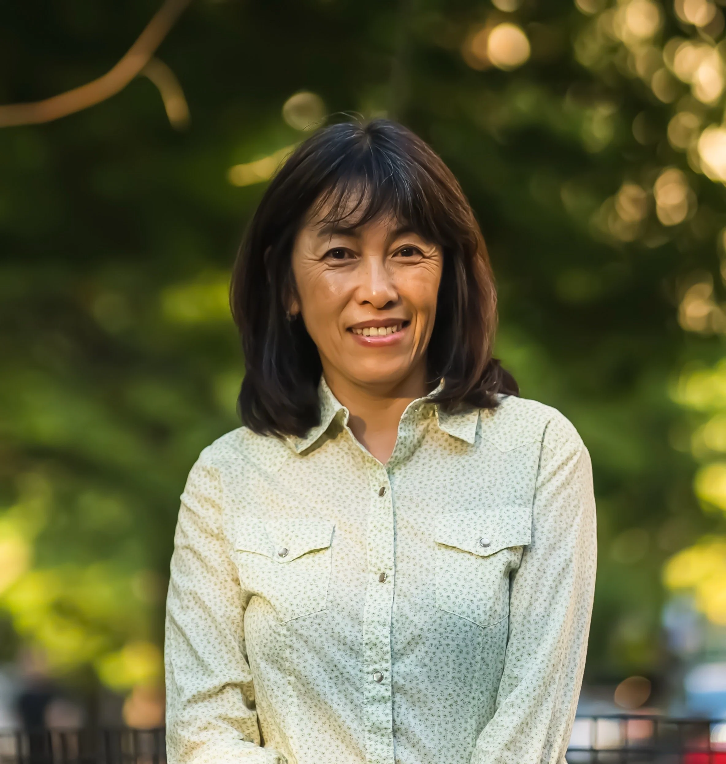 An Asian woman with dark brown hair standing outside, near trees, wearing a light green, patterned shirt. Photo by John McCormick.