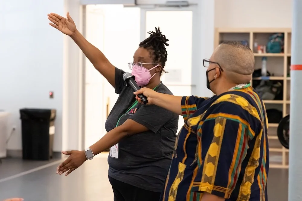 JJ, a disabled person, wears glasses, a face mask, and a colorful shirt, while holding out a microphone with their right hand in front of Kayla, a disabled Black woman wearing glasses, a face mask, a gray t-shirt, and her hair in a braided, top bun.