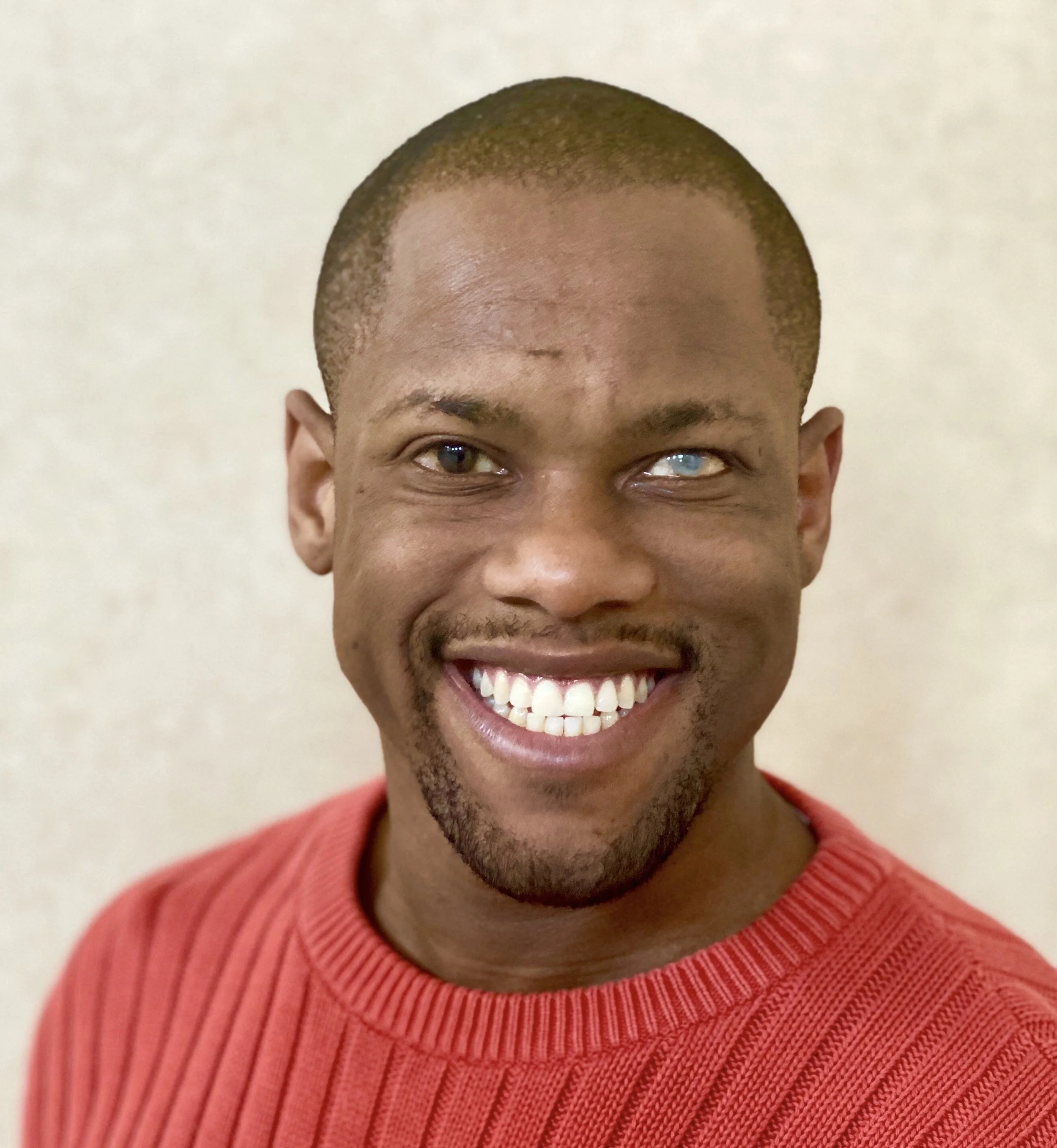 A smiling man wearing a red  sweater. His hair is cropped, and he has a neatly trimmed mustache and goatee. One eye appears clear while the other appears cloudy, suggesting visual impairment.