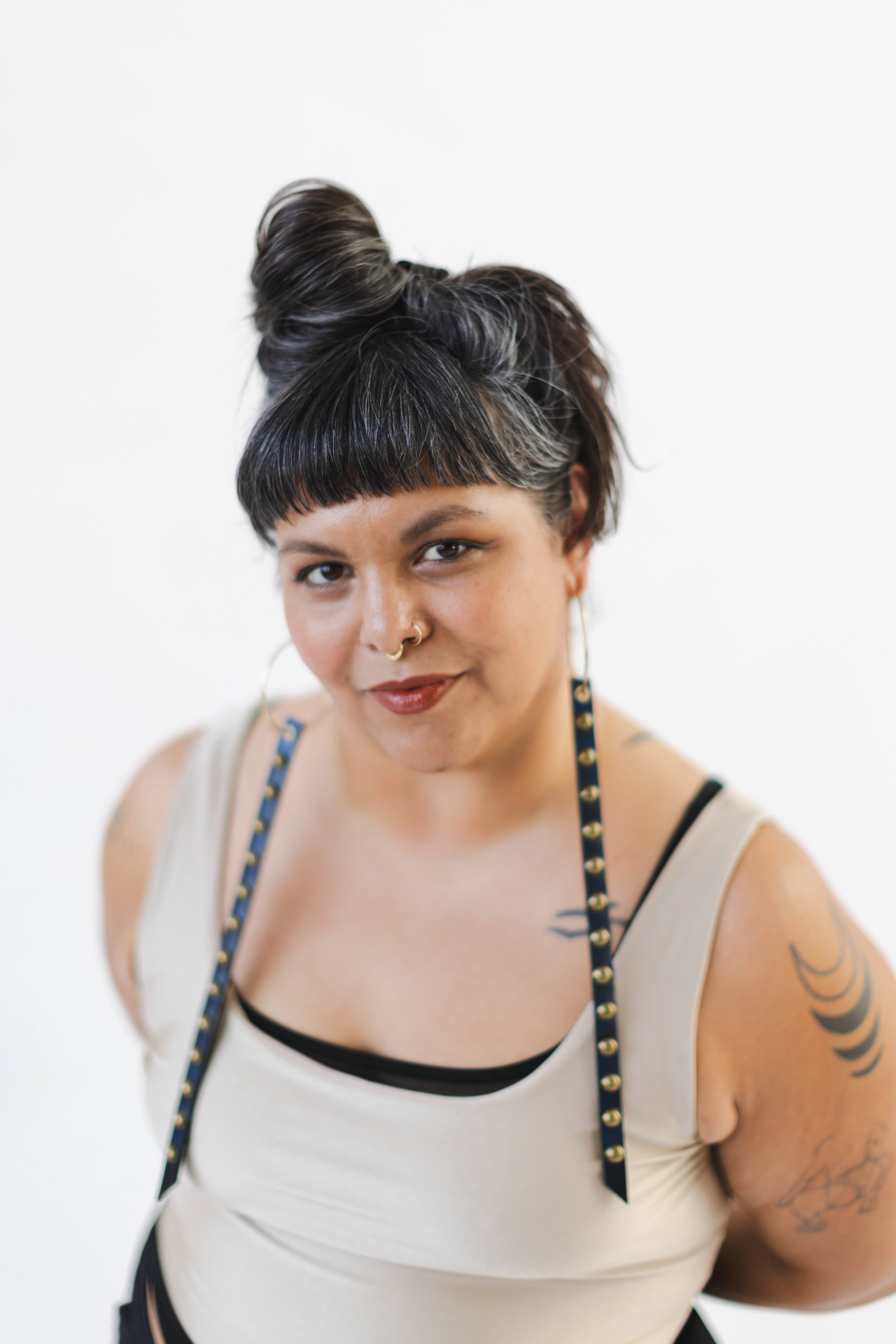 A non-binary femme with cinnamon skin. Their hair is in a bun with dark brown bangs streaked with silver and their skin is artfully tattooed. They wear nose piercings. statement earrings, a silver tank, and red lipstick. Photo by Isabel Fajardo.e