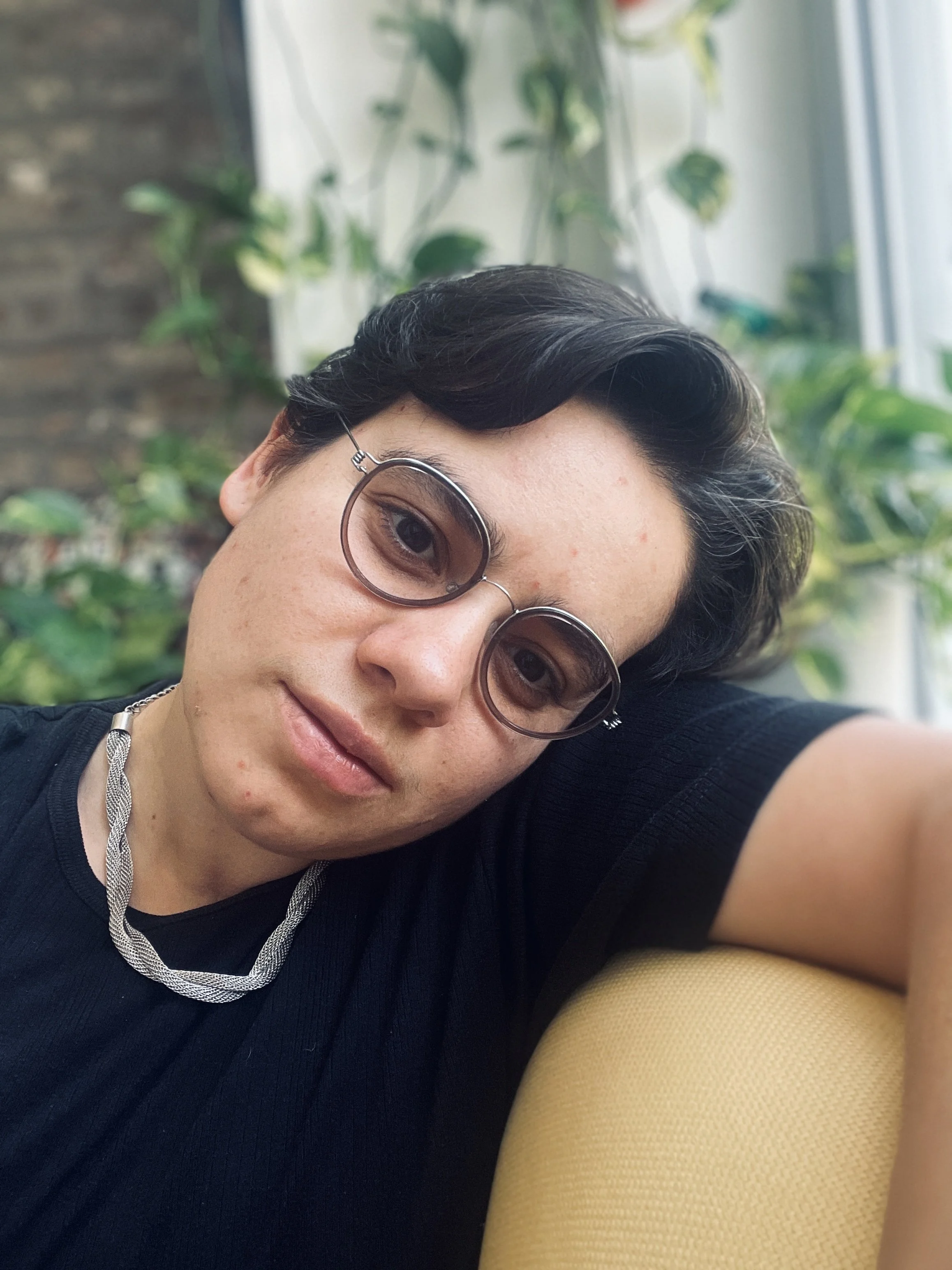 A Puerto Rican, nonbinary person with light skin and short, dark brown hair wearing black t-shirt and round glasses with violet tinted lenses. Ariana rests their head on their arms with sunlit brick and greenery behind. Photo courtesy of artist.