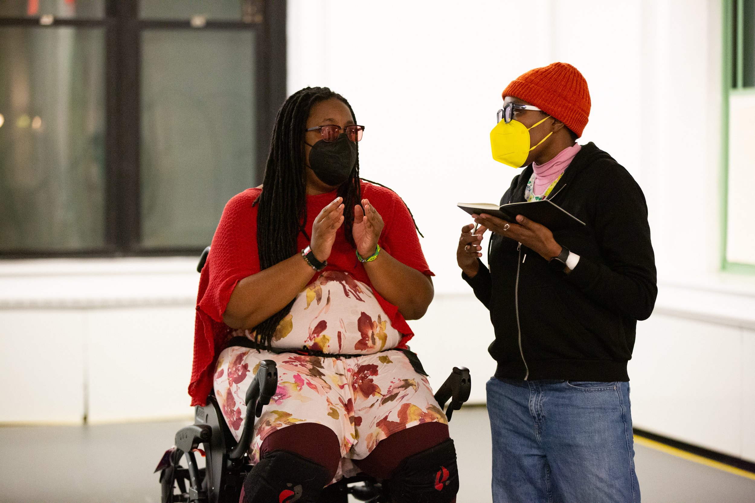 Two Black individuals wearing face masks are conversing. One of them is in a wheelchair wearing a dress, the other is wearing a beanie and holds an open book.