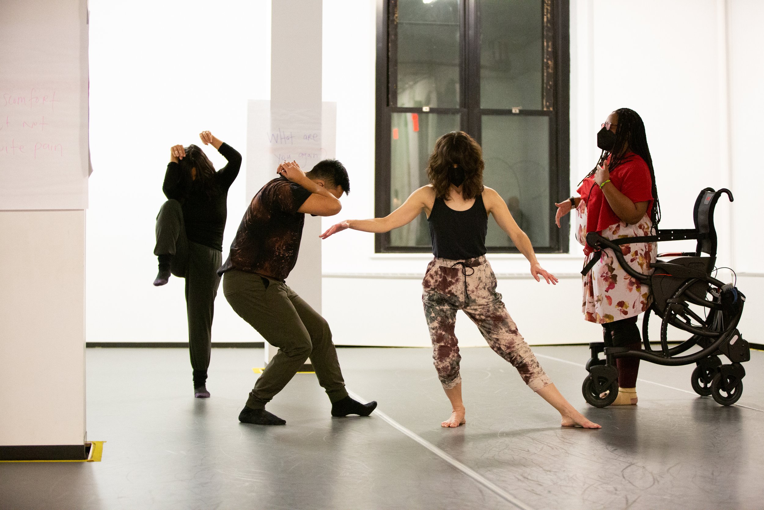 A group of people rehearsing a dance performance. They are seen from behind, and a wheelchair is visible.