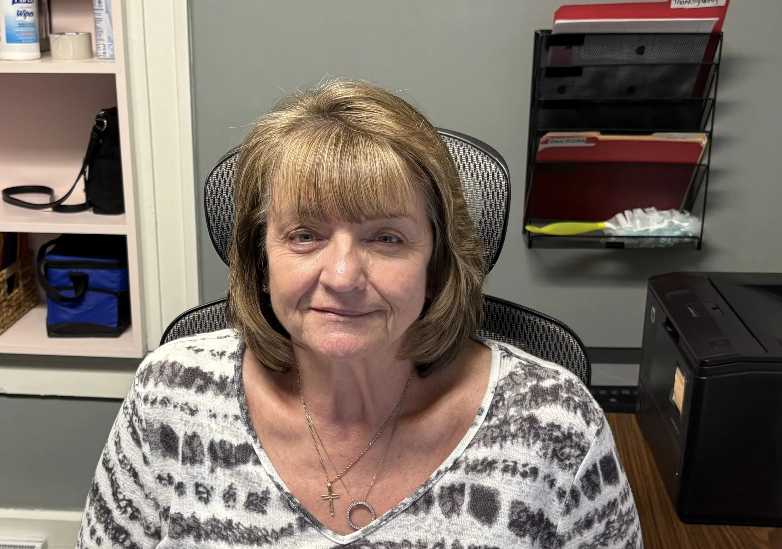 A woman with shoulder-length blonde hair sitting in an office chair, smiling, wearing a gray zip-up hoodie over a gray shirt.