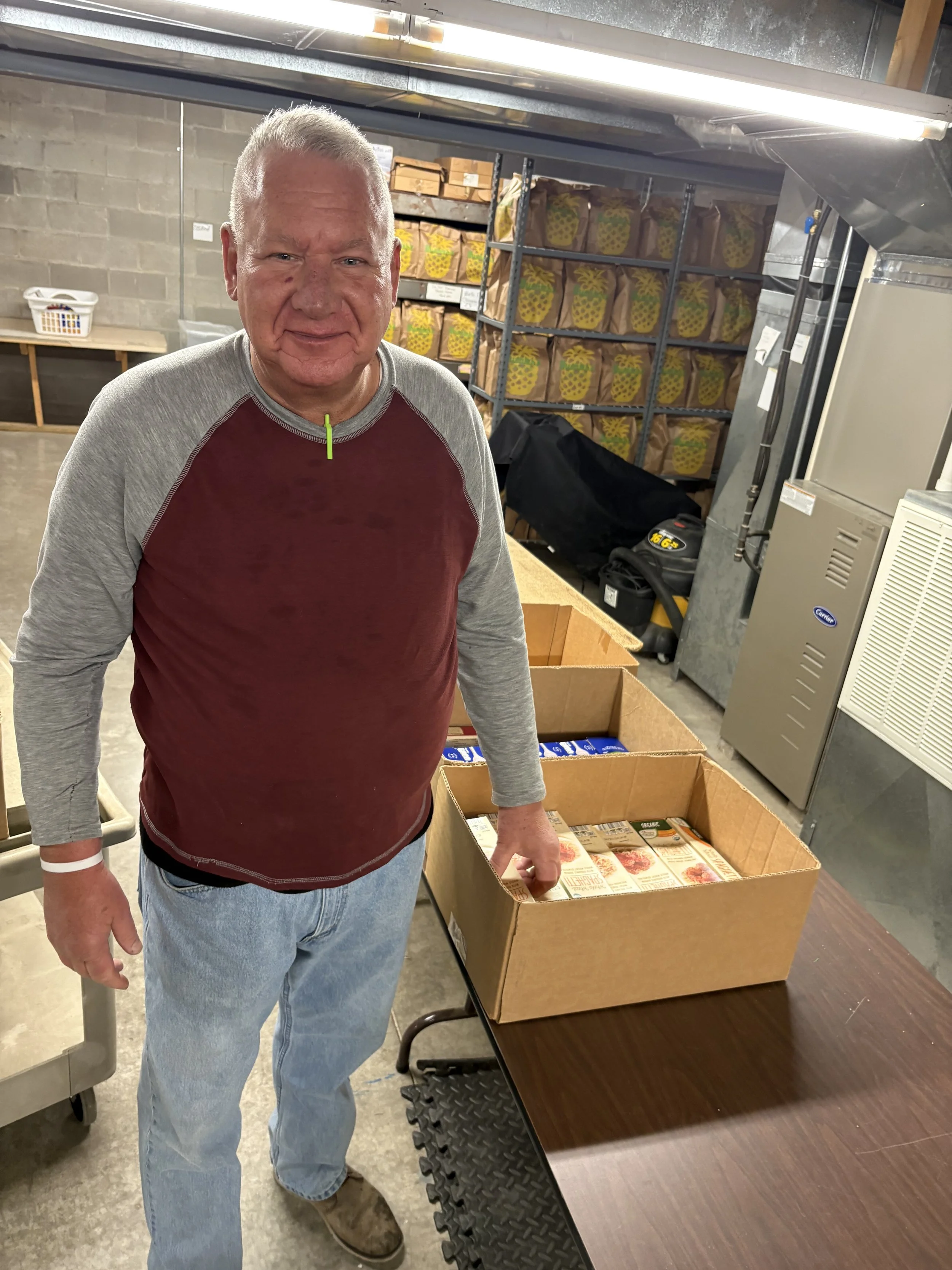 A man standing next to a table with open cardboard boxes filled with packaged food items, in an industrial setting with shelves of pineapples in the background.