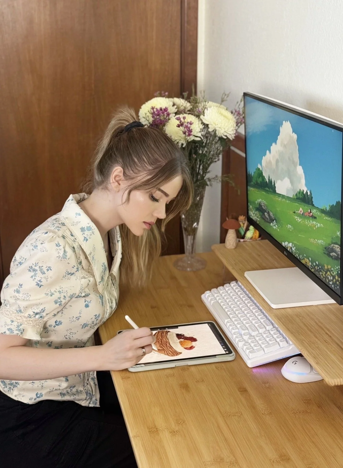 A woman with long brown hair tied in a ponytail, wearing a cream-colored blouse with blue floral patterns, is sitting at a wooden desk and drawing on a tablet with a stylus. There is a computer monitor on the desk displaying a scenic landscape with green hills, rocks, trees, and a blue sky with clouds. In the background, a glass vase with white and purple flowers is visible.